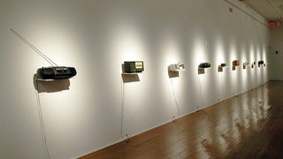 Ausschnitt der Soundperformance "Deterritoriale Schlingen" von Thom Kubli und Sven Mann, aus dem Jahr 2004. Zu sehen sind diverse Radioempfangsgeräte, stehend auf Regalbretten, die an der Wand montiert sind.
