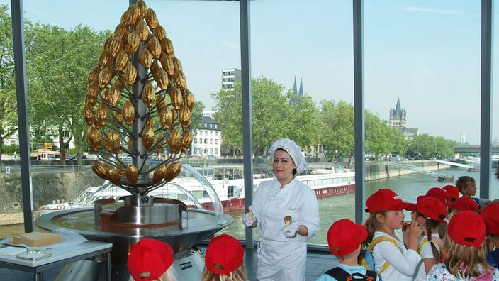 Kinder und eine Museumsangestellte stehen am Schokoladenbrunnen, im Hintergrund das Rheinufer 