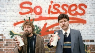 Jürgen von der Lippe und Gerd Dudenhöfer stehen mit Spraydosen in der Hand vor einer Hauswand, auf der in roter Farbe der Schriftzug "So isses" prangt (1984)