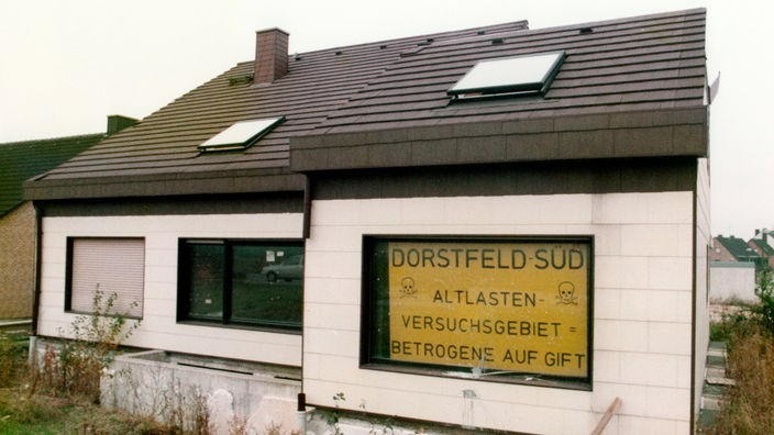 Ein Wohnhaus in der Dortmunder Neubau-Siedlung in Dorstfeld-Süd. Im Fenster ein Schild mit Schriftzug "Dorstfeld-Süd; Altlasten-Versuchsgebiet = Betrogene auf Gift" (1985).