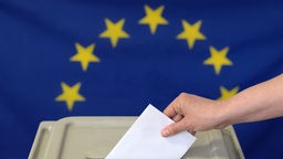 Eine Hand steckt einen Umschlag in eine Wahlurne vor einer Europafahne.
