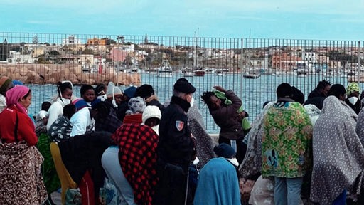Flüchtlinge stehen vor einem Grenzzaun.
