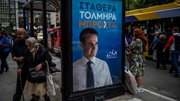 Vor den Parlamentswahlen gehen Menschen neben einem Wahlkampfplakat mit einem Porträt des Ministerpräsidenten Mitsotakis an einem Busbahnhof vorbei, Athen,  11.05.2023. 