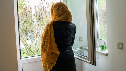 Symbolbild: Eine in ihr Kopftuch eingehüllte Muslima steht an einem geöffneten Fenster.