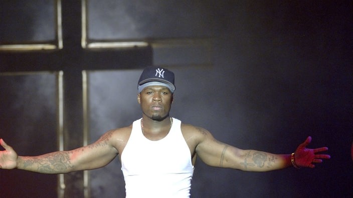 Der us-amerikanische Rapper 50 Cent steht auf einer Bühne in verheißungsvoller Pose vor einem großen Kreuz.