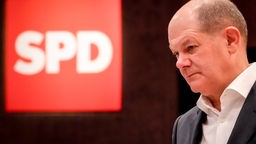 Olaz Scholz mit nachdenklichem Gesichtsausdruck neben einem Logo der SPD
