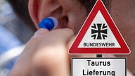 Symbolbild: Ein Warnschild "Bundeswehr" "Taurus-Lieferung" vor einem Mensch mit Kopfhörer