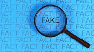Eine Lupe hebt von einer mit "Fact" beschriebenen Seite das Wort "Fake" hervor.