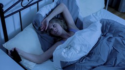 Frau liegt im Bett und schaut müde auf die Uhr. Symbolbild Schlaflosigkeit