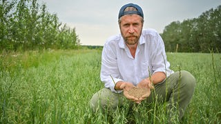 Landwirt Benedikt Bösel hock in legren Outfit auf einem Acker und hält Erde in die Kamera.