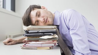 Ein Mann im Arbeits-Hemd legt den Kopf auf einen Stapel mit Unterlagen und träumt.