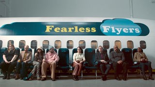 Mehrere Menschen Sitzen in einer Reihe auf stühlen vor einem Flugzeug.