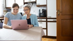 Großmutter und Enkeltochter sitzen an einem Küchentisch gemeinsam vor dem Laptop.