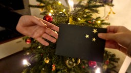 Symbolbild Gutscheine verschenken: Zwei Hände halten einen Umschlag, im Hintergrund ein Weihnachtsbaum