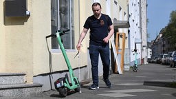 Der blinde Reporter Kevin Barth mit einem Langstock auf dem Gehweg, den ein E-Scooter blockiert