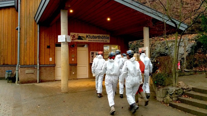 Mehrere Besucher in weißen Schutzanzügen betreten den Eingangsbereich vor der Kluterhöhle. 