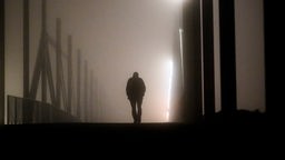 Der Umriss eines Mannes der mit gesunkenem Haupt mitten auf einer Brücke entlanggeht.