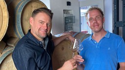 Max Giermann und Daniel Schlipf stoßen vor Weinfässern mit Weingläsern an