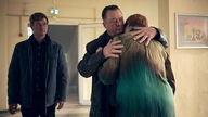 Szene aus „Polizeiruf 110: Der Dicke liebt“: Henry Koitzsch (Peter Kurth) umarmt Monika Hollig (Susanne Böwe) in einer Schule; im Hintergund Michael Lehmann.