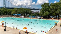 Mehrere Menschen Baden in einem Schwimmbad in New York vor stadttypischer Kulisse.