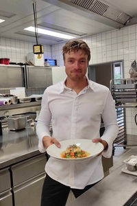 Sternekoch Torben Schuster steht in seiner Küche und hält einen Teller Papaya-Salat mit Seeteufel