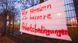 Auf einem Transparent an einem Zaun steht "Wir streiken für bessere Arbeitsbedingungen"