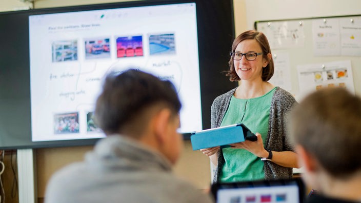 Eine Lehrerin steht mit einem iPad vor einer digitalen Schultafel im Englischunterricht einer 5. Klasse