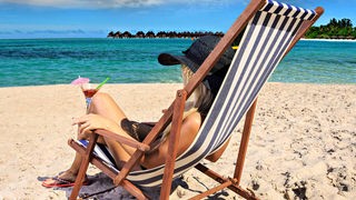 Eine junge Frau mit Sonnenhut sitzt auf einem gestreiften Liegestuihl an einem Strand auf den Malediven und blickt auf das Meer. In der Hand hält sie einen Coctail.
