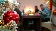 Im Vordergrund hängt eine Christbaumkugel, im Hintergrund sitzt eine Familie beim Weihnachtsessen.