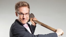 Der Kabarettist Benjamin Eisenberg posiert mit Vorschlaghammer vor grauem Hintergrund, Foto: Dominik Sutor