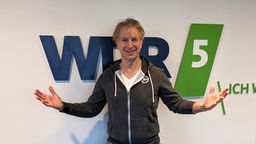 Schauspieler und Moderator Ingolf Lück steht vor dem WDR-5-Logo und breitet lächelnd die Arme aus