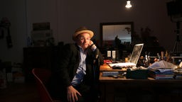Der Kabarettist Thomas Pigor sitzt an seinem Schreibtisch vor dem Laptop und schaut in die Kamera