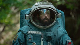 Adam Sandler als Kosmonaut Jakub in einem großen Raumanzug inmitten eines grünen Waldes.