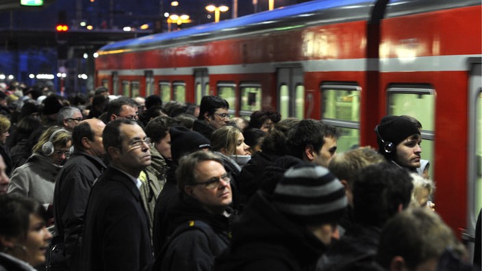 Fahrgäste der Bahn stehen an einem vollen Gleis, während eine S-Bahn einfährt.