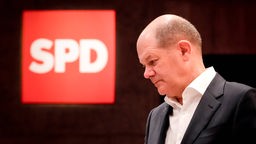 Olaf Scholz mit gesenktem Haupt. über ihm das leuchtend rote Logo der SPD zu sehen.