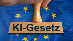 Ein symbolischer Holzstempel mit der Aufschrift "KI-Gesetz", gehalten von einer Hand im Anschnitt vor einer wehenden EU-Flagge im Hintergrund. 