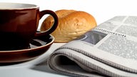 Eine Sonntagszeitung liegt neben einer Tasse Kaffee und einem Brötchen. 
