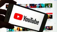 Das Logo des Video-Portals YouTube wird auf dem Display eines Smartphones angezeigt. Im Hintergrund ist auf einem Bildschirm die YouTube Homepage zu sehen