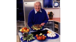Robert Atkins, ein schlanker Mann mit weißen Haaren steht in einer KÜche und hält freundlich lächelnd einen Teller mit viel Fleisch in die Kamera, Aufnahme aus dem Jahr 1999