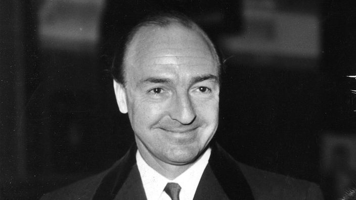 John Dennis Profumo, ein Mann mit hoher Stirn im dunklen Anzug lächelt zufrieden. Schwarz-weiß Aufnahme aus dem Jahr 1960