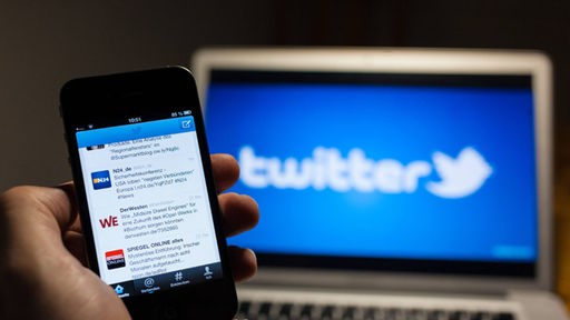 Ein Nutzer hält ein Smartphone, im Hintergrund zeigt ein Bildschirm das Twitter-Logo
