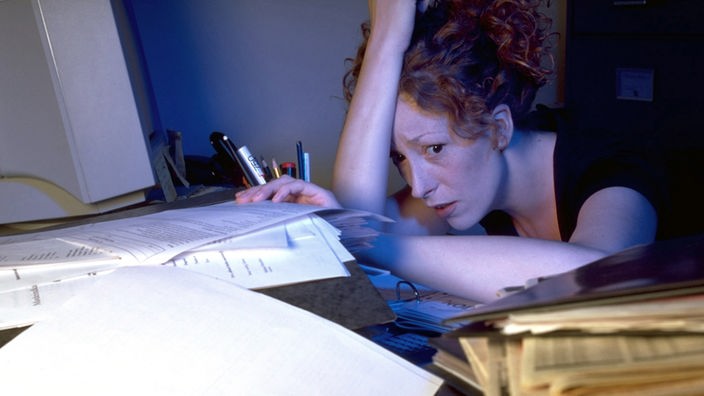 Eine Frau stützt sich am Schreibtisch ab, vor ihr ein Haufen von Arbeit