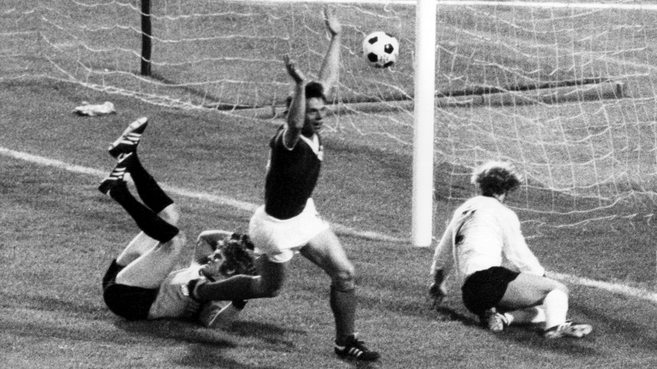 Jürgen Sparwasser reißt jubelnd die Hände hoch, gegenerischer Torwart und Spieler liegen am Boden, der Ball fliegt ins Netz