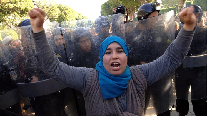 Eine Frau mit Kopftuch demonstriert, im Hintergrund Militär
