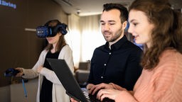 Junge Frau mit VR-Brille, junger Mann und junge Frau schauen in Laptop