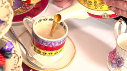 Das Bild zeigt eine bunte Tasse, die mit schwarzem Tee mit Milch befüllt wird