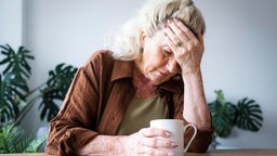 Eine ältere Dame sitzt gestresst am Tisch und schaut in ihre Tasse.