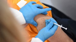 Zwei Hände mit blauen Handschuhen setzen eine Impfspritze in einen Oberarm