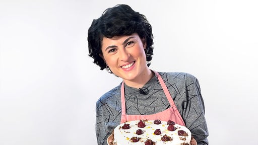 Konditormeisterin Theresa Knipschild mit einer Torte in Händen und einer rosafarbenen Weste vor weißem Hintergrund.