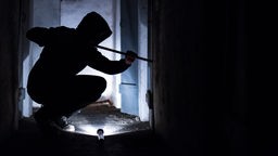 Ein Mann hebelt mit einem Brecheisen eine Tür im Keller eines Wohnhauses auf.